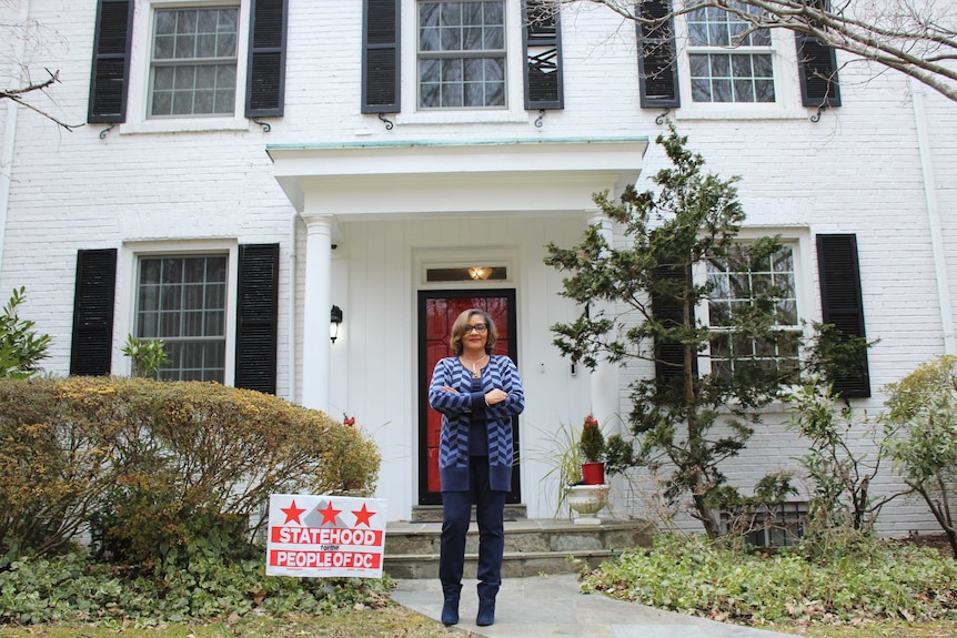 A woman in a blue chevron print cardigan standing outside a DC home with a 'statehood for people of DC' sign in the grass