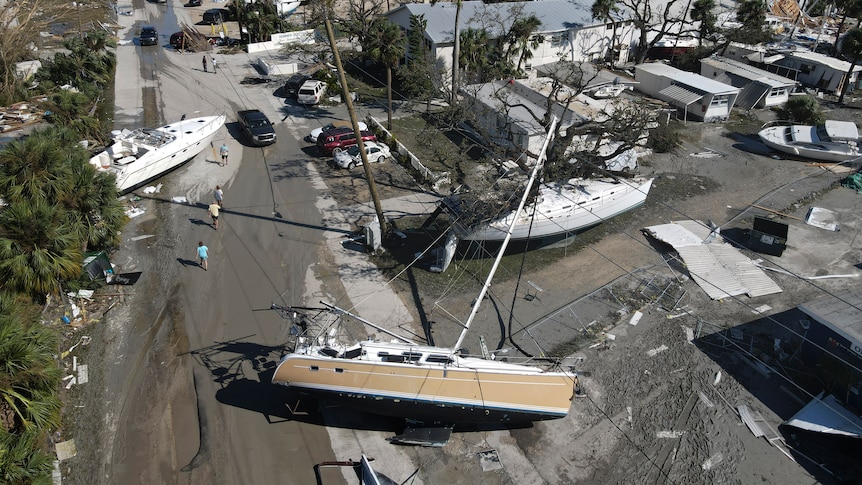 Damaged boats and large structures lie strewn between mobile homes on a seaside concrete and grass area.