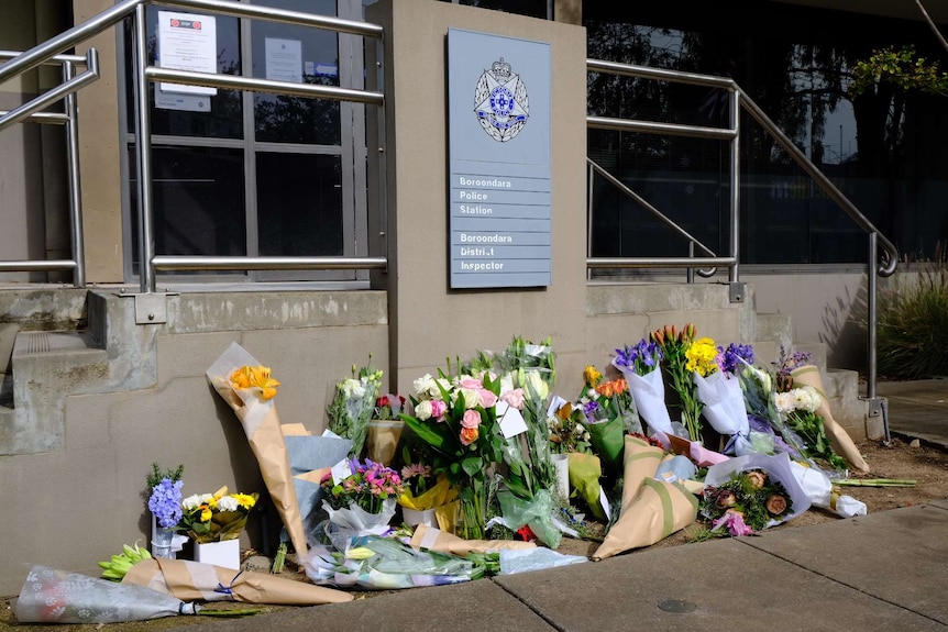 Bouquets of flowers are laid outside a police station on a sunny day.
