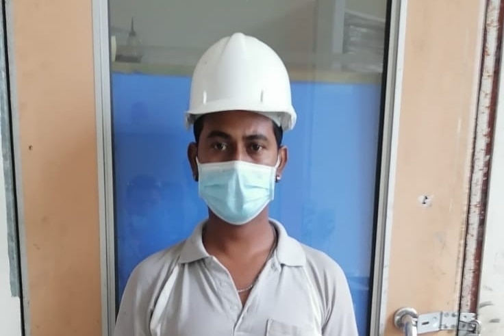 A young Bangladeshi man wearing a helmet and face mask standes next to a door frame in factory.