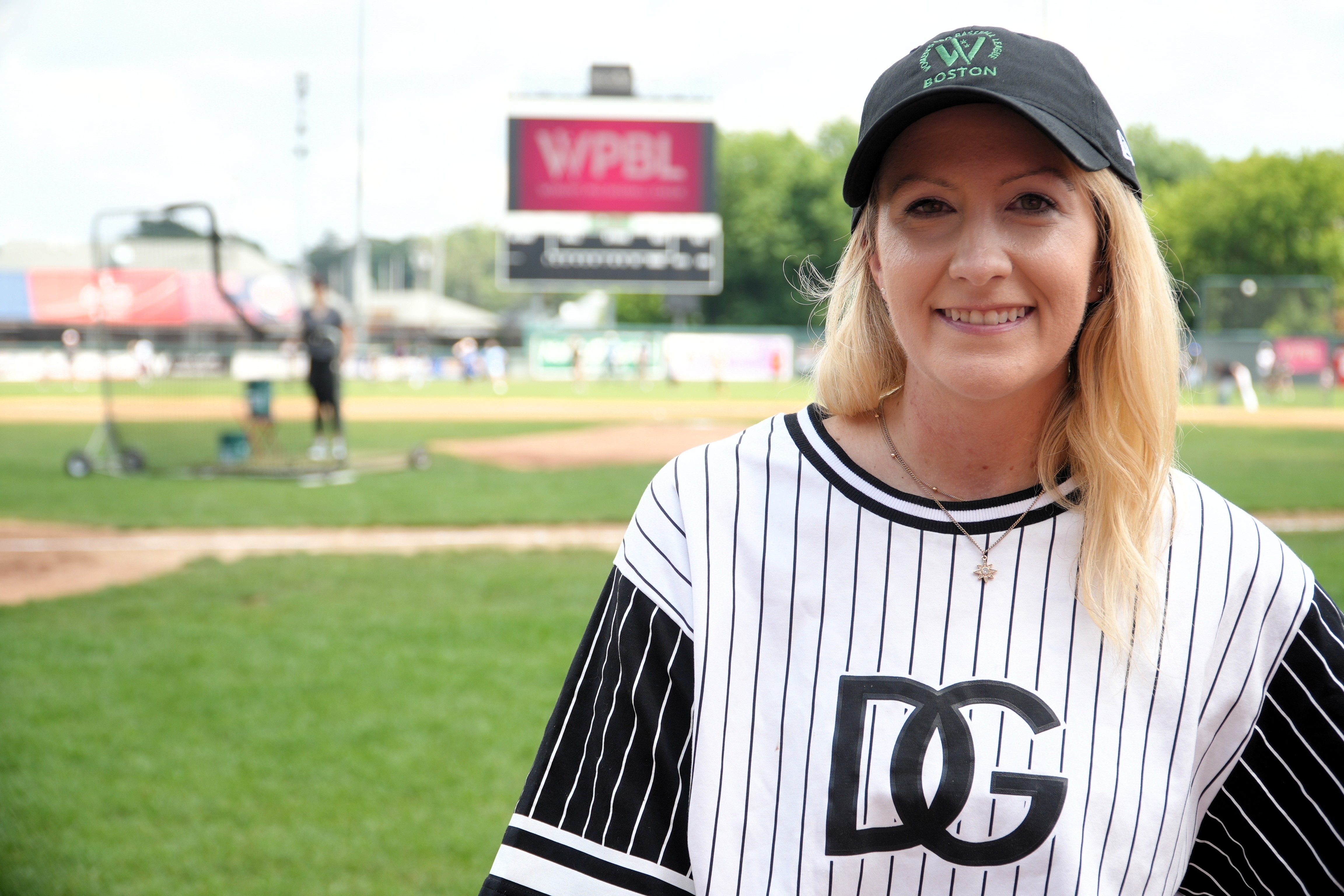A woman with shoulder length blond hair wears a baseball cap and smiles