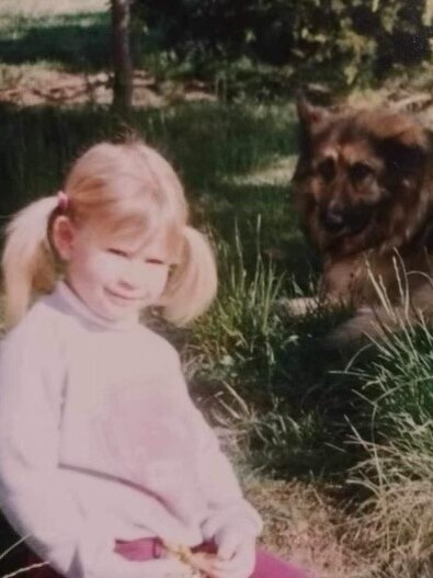 A young girl with two blonde pigtails smiles, sitting on the grass next to a German Shephard. 