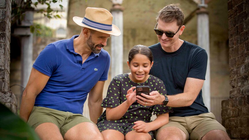 Two men sit with their daughter in a park while she looks at the screen of a mobile phone.
