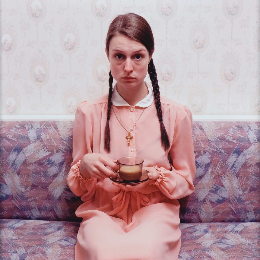 A woman with plaits in her hair and a crucifix necklace stares at the camera while holding a cup of tea