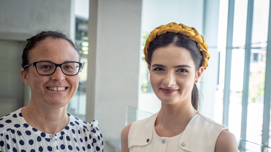 Two smiling women face the camera in the foyer of ABC Perth - one wears glasses, the other has a yellow headband