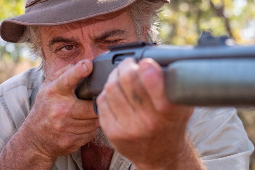 Man with a hat closes one eye and looks down the barrel of a rifle