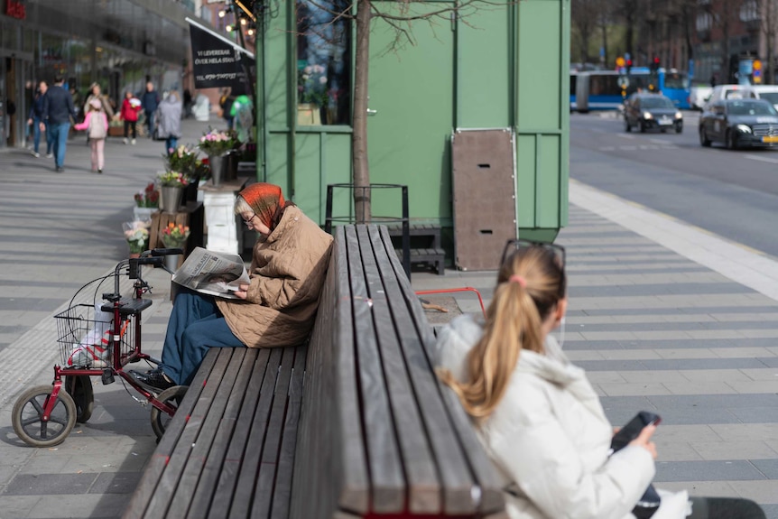 An elderly woman sits on a bench reading a newspaper in Stockholm.