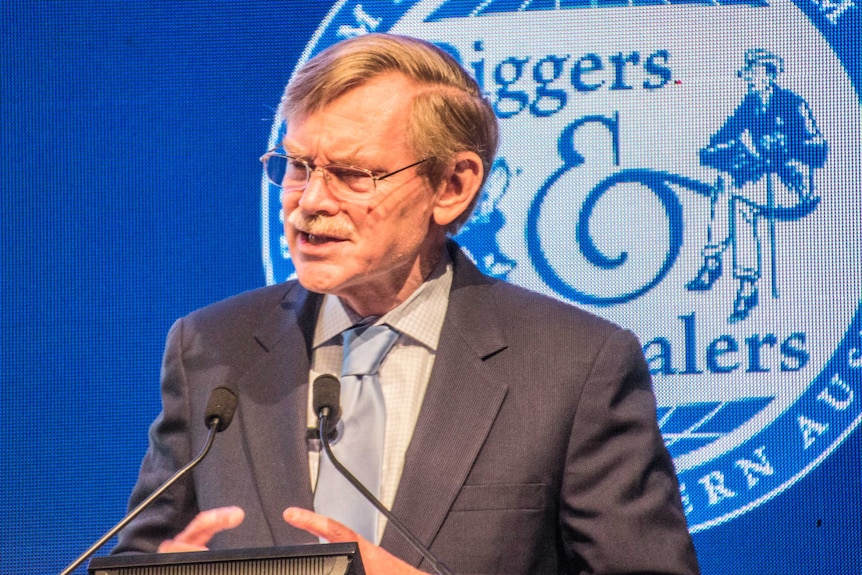 Man in suit speaks into microphones at a lectern with Diggersd and Dealers logo on screen behind him.