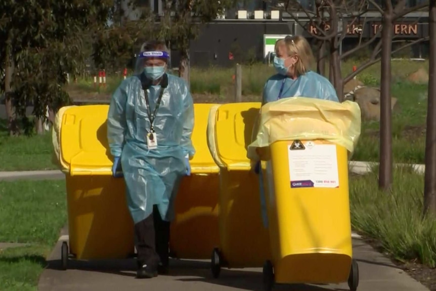 A man and a women wearing protective equipment wheeling four large, yellow hazardous materials bins.