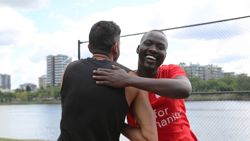 A Sudanese-Australian man wearing a red cross t-shirt is smiling broadly and clapping another man on the back
