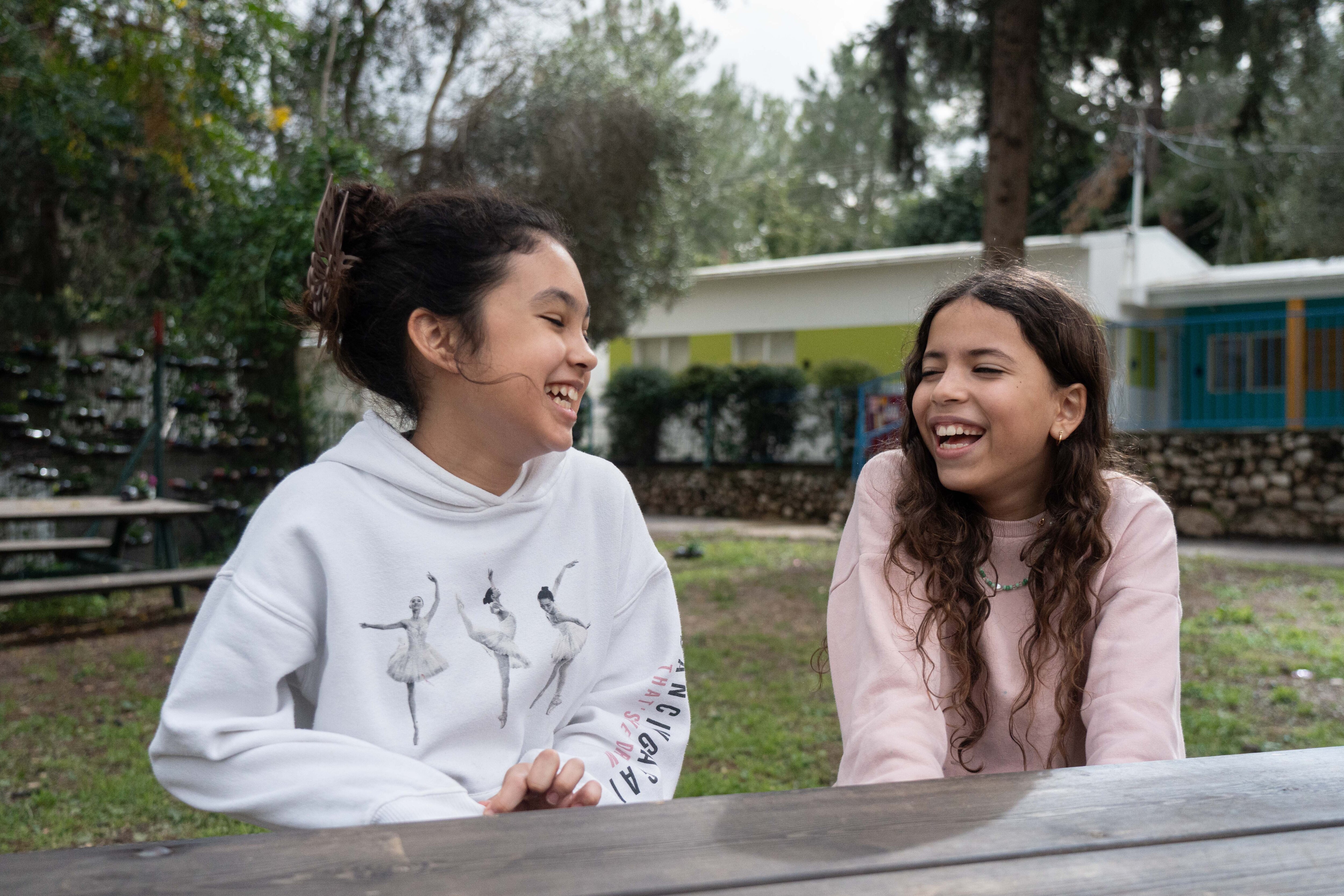 Two young girls sitting at an outdoor bench laughing together.