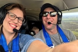 A man and woman with headsets on smile in a plane cockpit with a small child wearin a headset sitting in a car seat behind them.