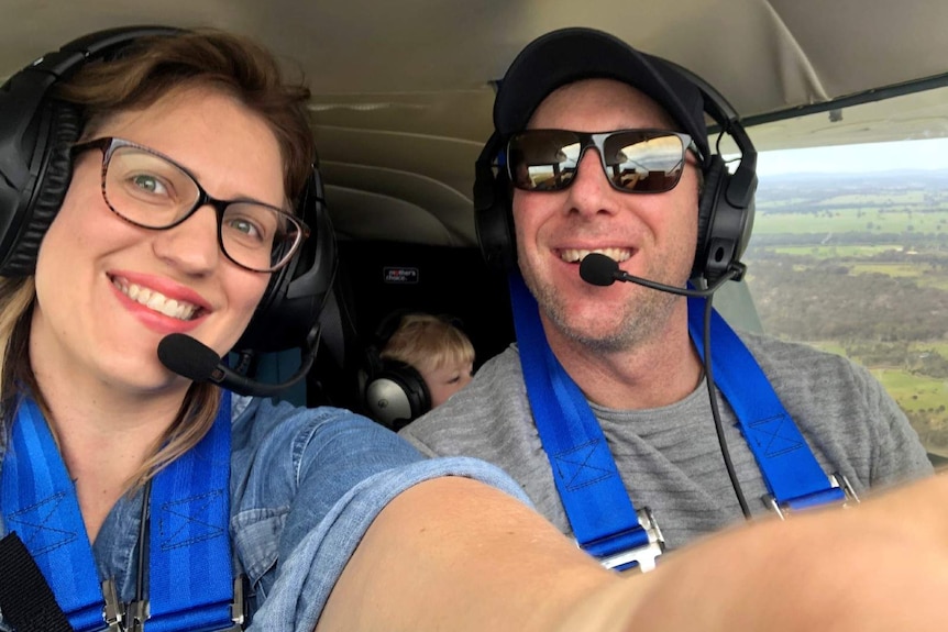 A man and woman with headsets on smile in a plane cockpit with a small child wearin a headset sitting in a car seat behind them.