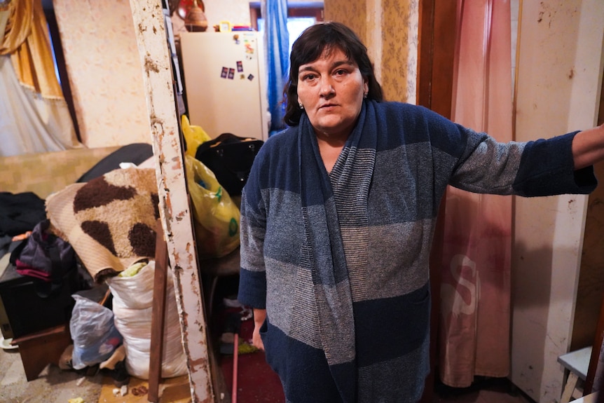A brunette woman wearing a warm jumper and jeans stands in a ruined home