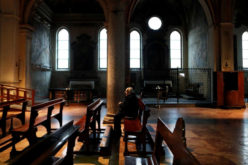An elderly man sits in a dimly lit and ornate church by himself.