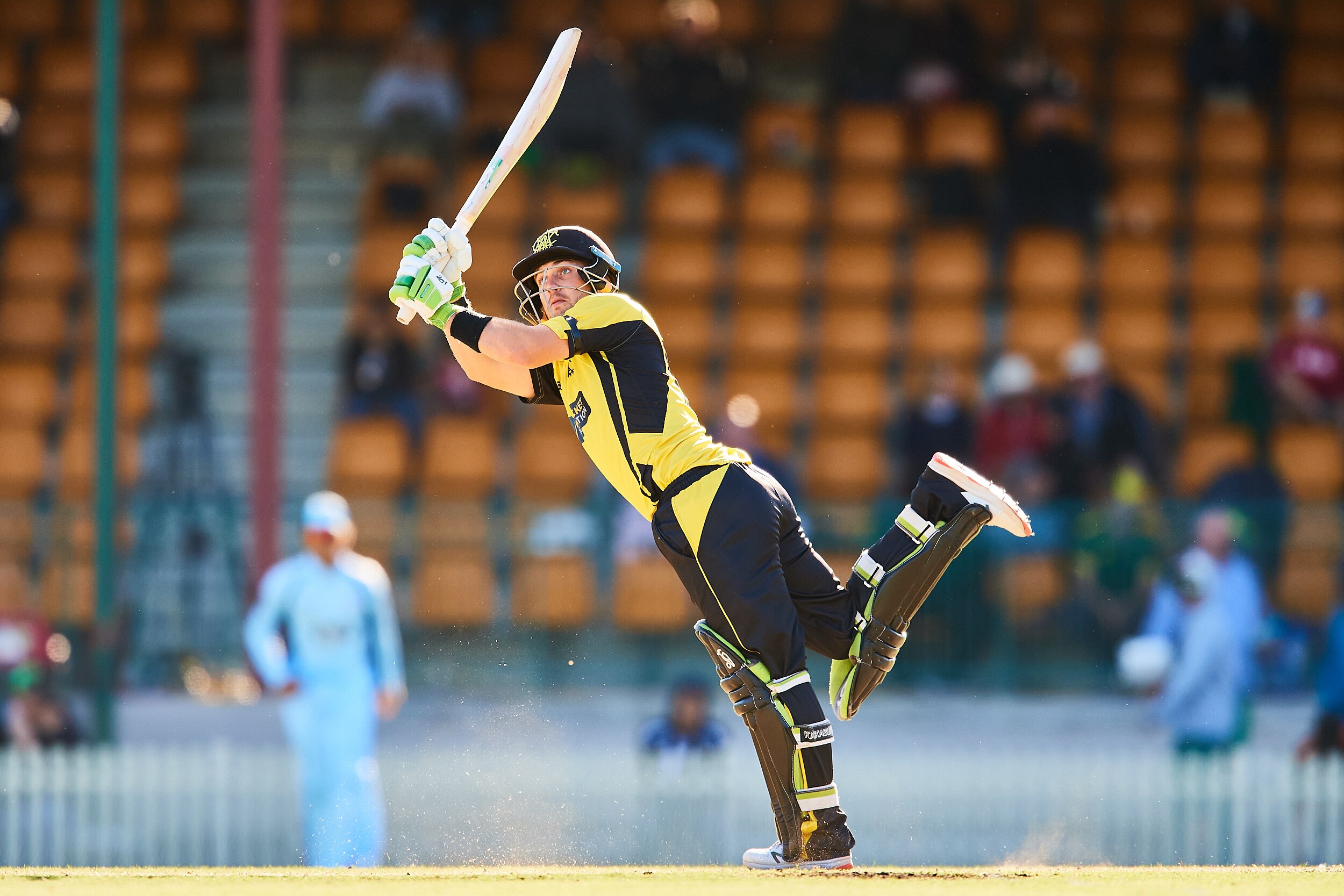 A batsman stands on one leg as he hits a lofted shot wide of mid-on.