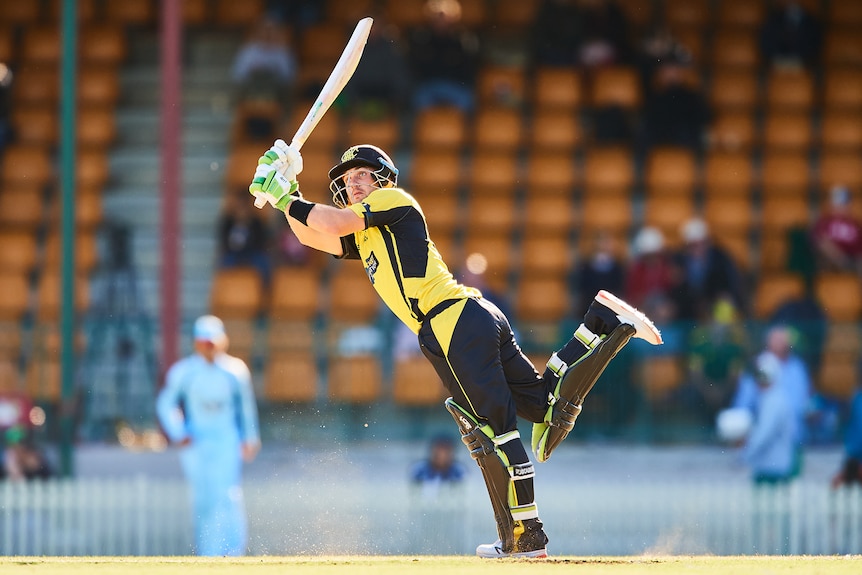 A batsman stands on one leg as he hits a lofted shot wide of mid-on.