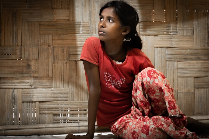 A girl in a red shirt and red patterned pants sits on the ground, staring off to her right.