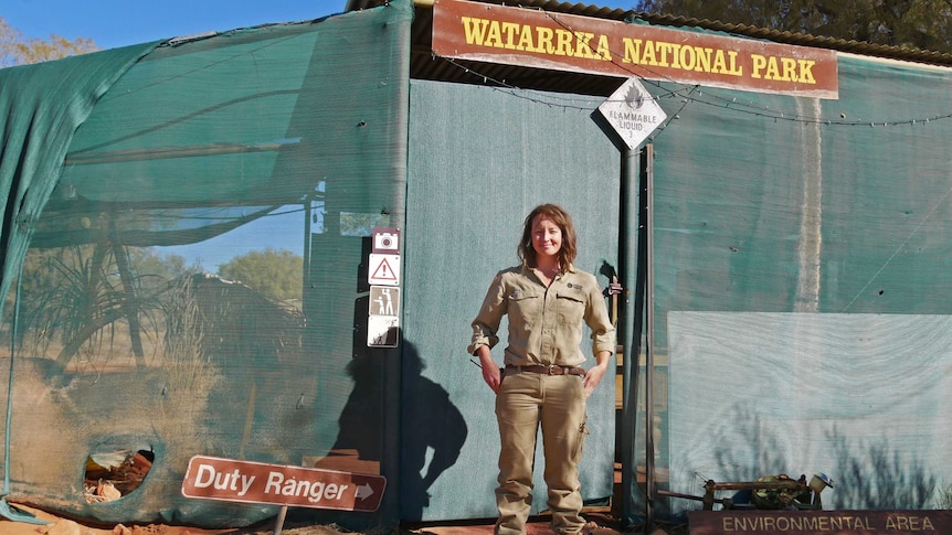 a woman in a park ranger uniform standing in front of a greenhouse with a Watarrka National Park sign.