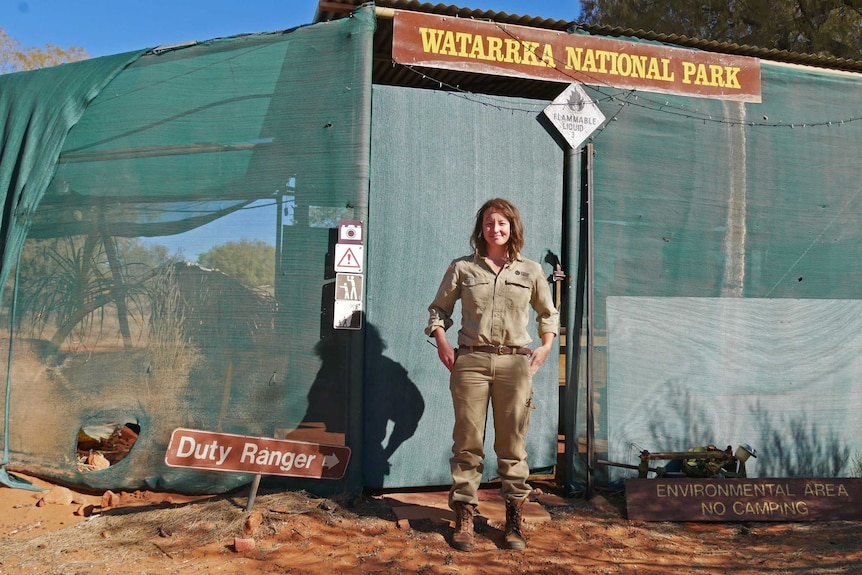 a woman in a park ranger uniform standing in front of a greenhouse with a Watarrka National Park sign.