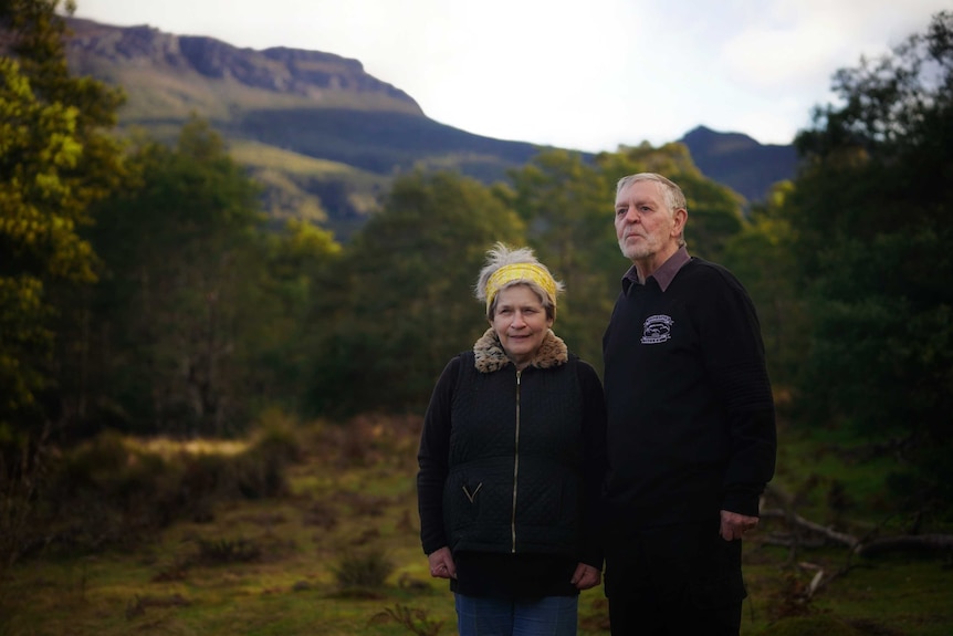 Pat and Len Doherty stand in a lush Tasmanian property, with mountains and escarpment behind them.
