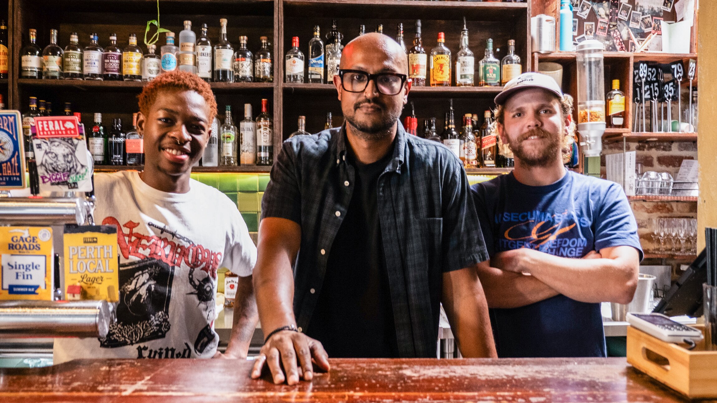 Three people stand behind a bar, the middle one with his hand on it.