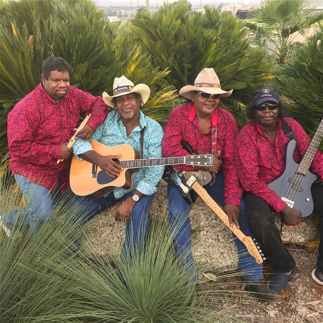Four men, two with guitars and hats and matching shirts sitting on rocks, smiling at the camera.