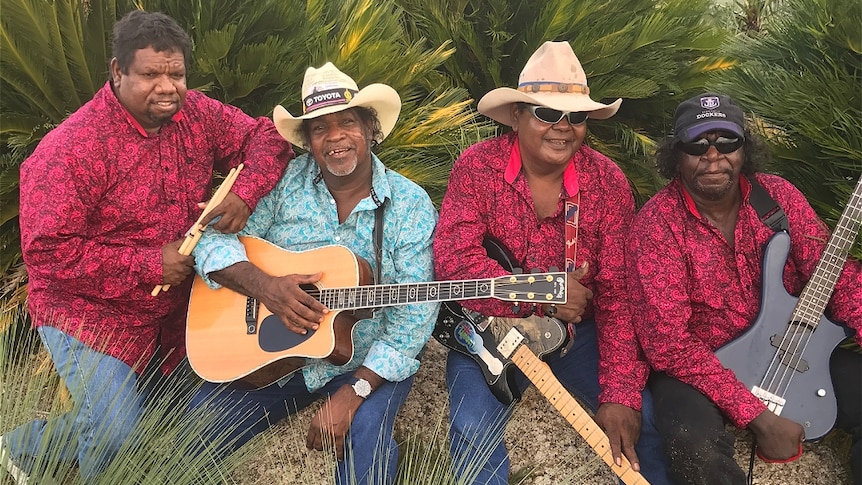 Four men, two with guitars and hats and matching shirts sitting on rocks, smiling at the camera.