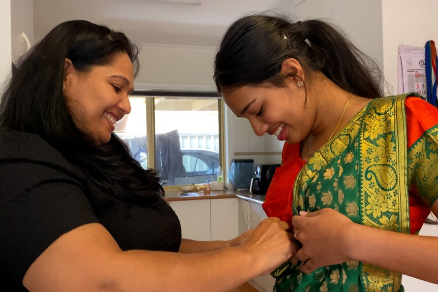 A brown woman is smiling as she pins a shiny green sash to her daughter's sari. It's a special moment.