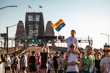 A person on another's shoulders holds a rainbow flag with a crowd visible in the background on the Sydney Harbour Bridge