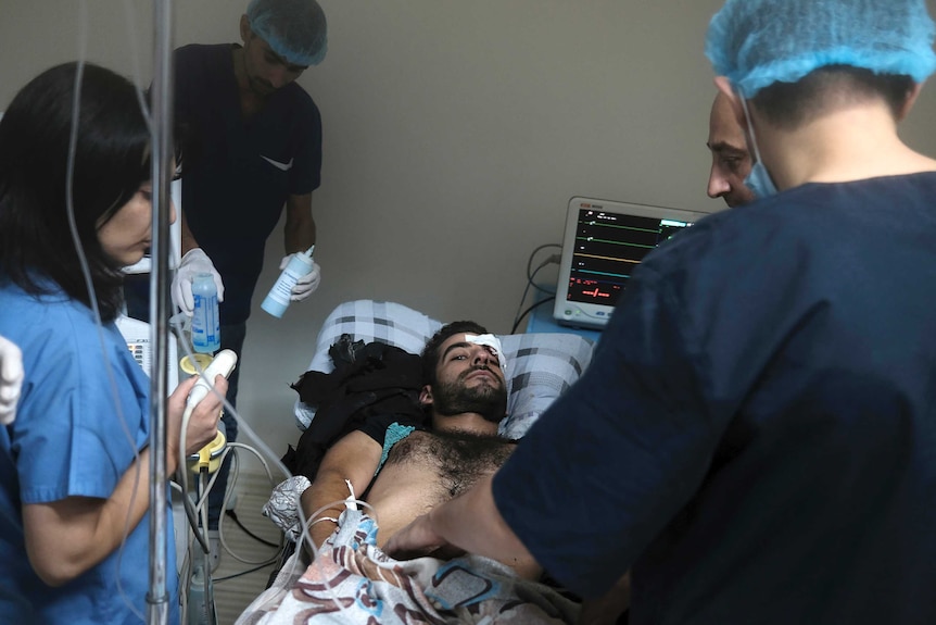 A man with dark hair and short beard stares at camera while lying on bed in hospital