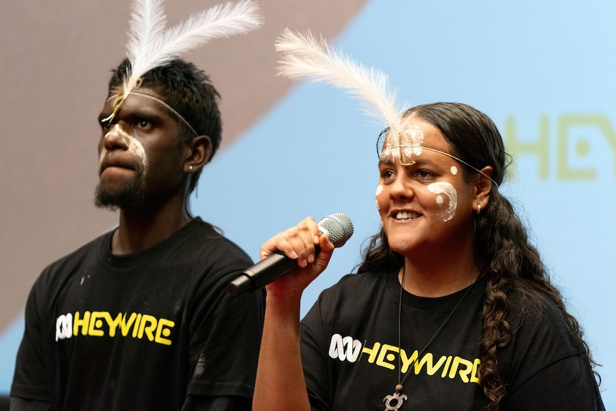 A young Indigenous man and woman with face paint and feathers wear black Heywire t-shirts.