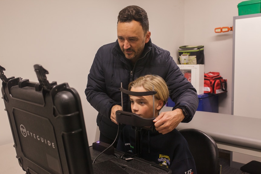 A man helps a boy use a machine that scans his eyes.