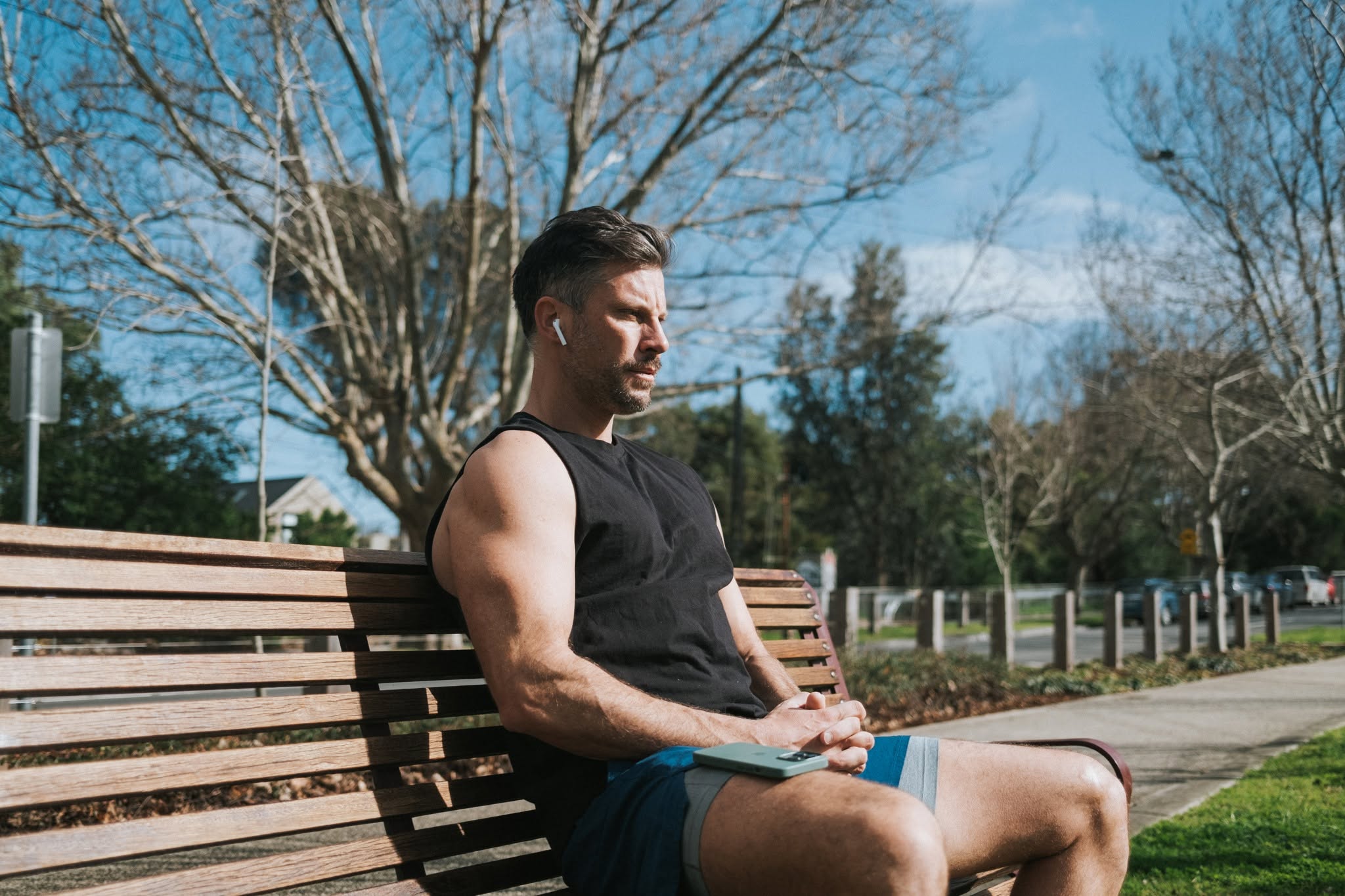 Muscular man in dark single, brightly-coloured shorts, on park bench with in-ear headphones