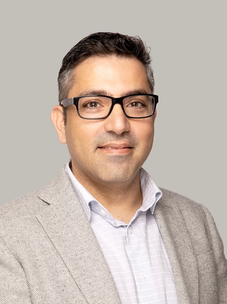 Dr Moksh Sethi wears black rimmed glasses, a white collared shirt and beige jacket.
