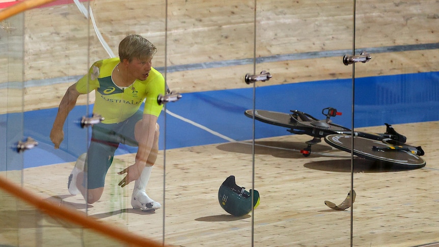 A shocked looking Australian cyclist kneels on the track as his pursuit bike lies in bits around him. 