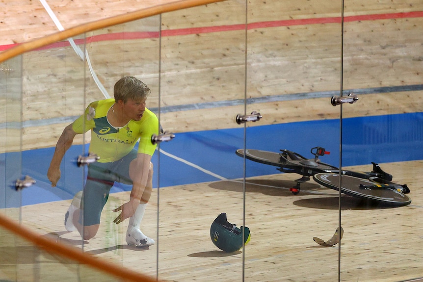 A shocked looking Australian cyclist kneels on the track as his pursuit bike lies in bits around him. 