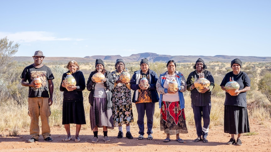 A man and seven women stand in a line, each holding colourful ceramic pots, with a desert backdrop.