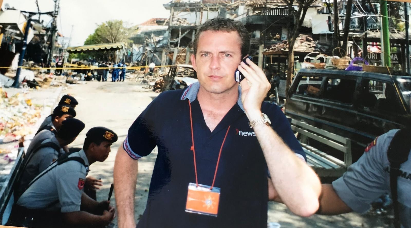 Young man in black polo short on phone with bomb damaged building in background