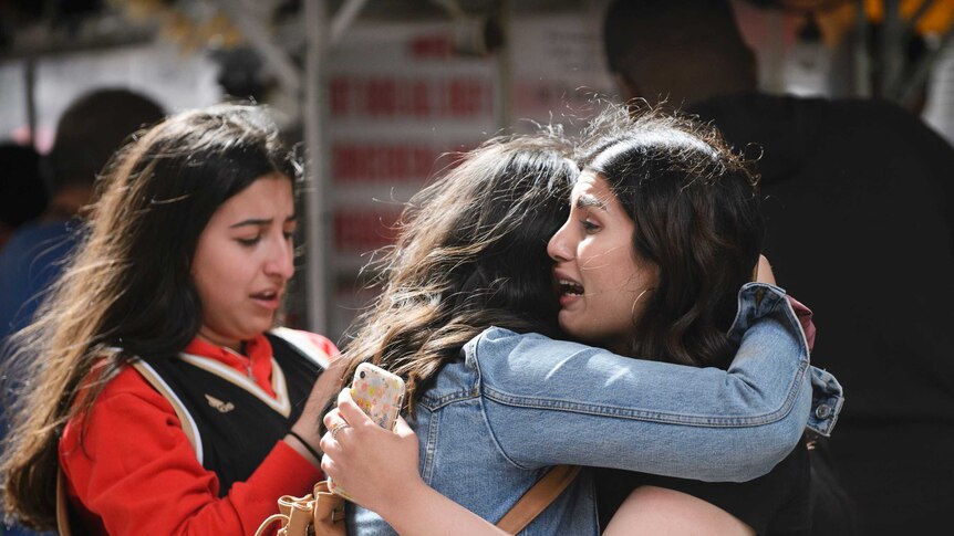 Two young women hug while another appears to be texting while crying behind them.