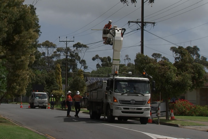 A white truck with a crane carrying a person wearing high-vis orange clothing with powerlines above and workers on the ground 