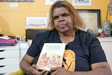 An elderly indigenous woman holds a book written called Tjanimako Tjukurpato featuring a scene from Aboriginal life