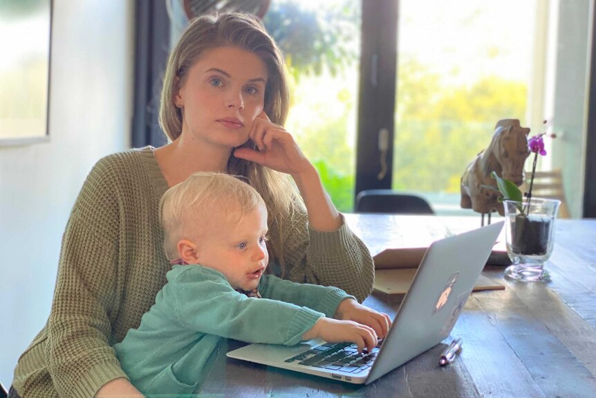 Dr Anna Samecki rests her head on a hand, sitting at a table in front of a laptop with her toddler daughter on her lap in Sydney