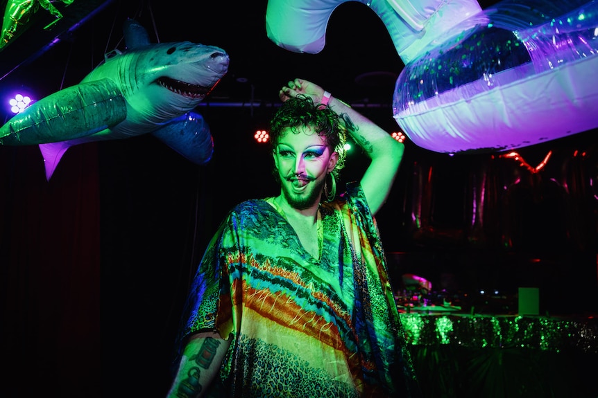 A drag queen performer dances on stage with inflatable sharks and toys behind them, washed in a green light. 