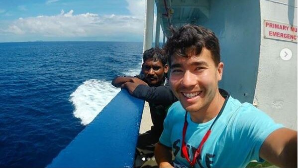 A man takes a selfie on a boat with another man leaning on the rail behind him.