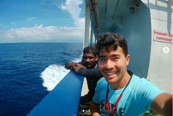 A man takes a selfie on a boat with another man leaning on the rail behind him.