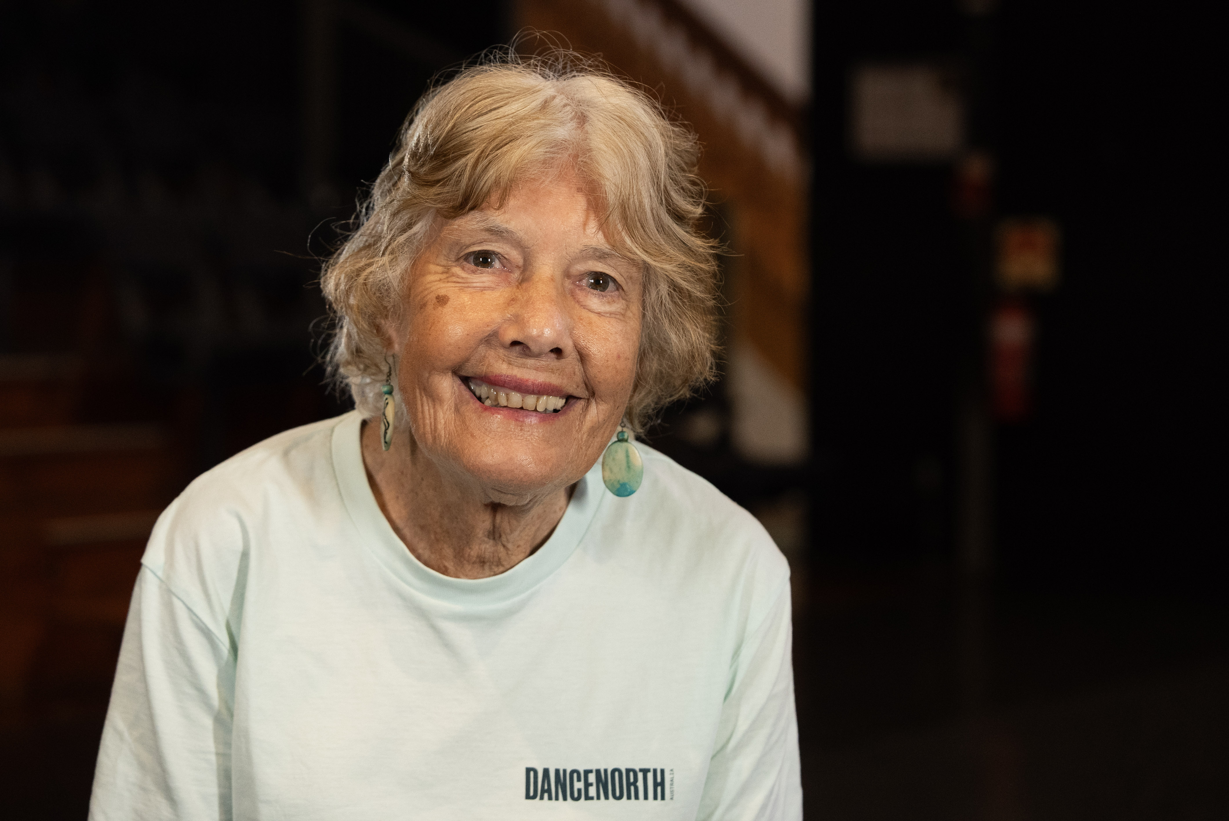 An older woman with medium length grey hair in a white shirt looks at the camera.