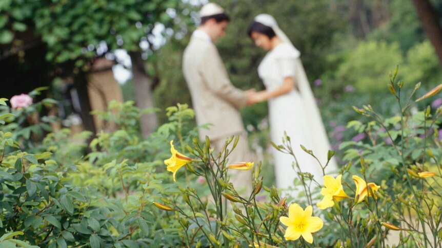 A Jewish couple hold hands in the background on their wedding day, while the camera focuses on a bright yellow flower.