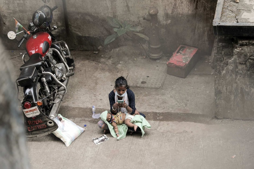 A girl looks at her phone with a baby sleeping in her lap next to a motorcycle.