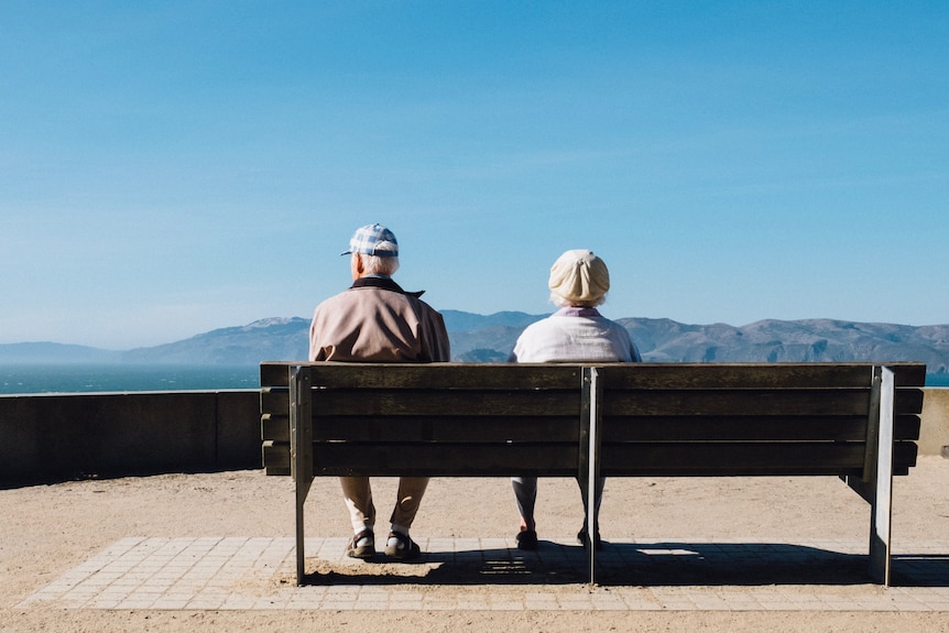 Older man and woman sit on bench looking out at landscape.